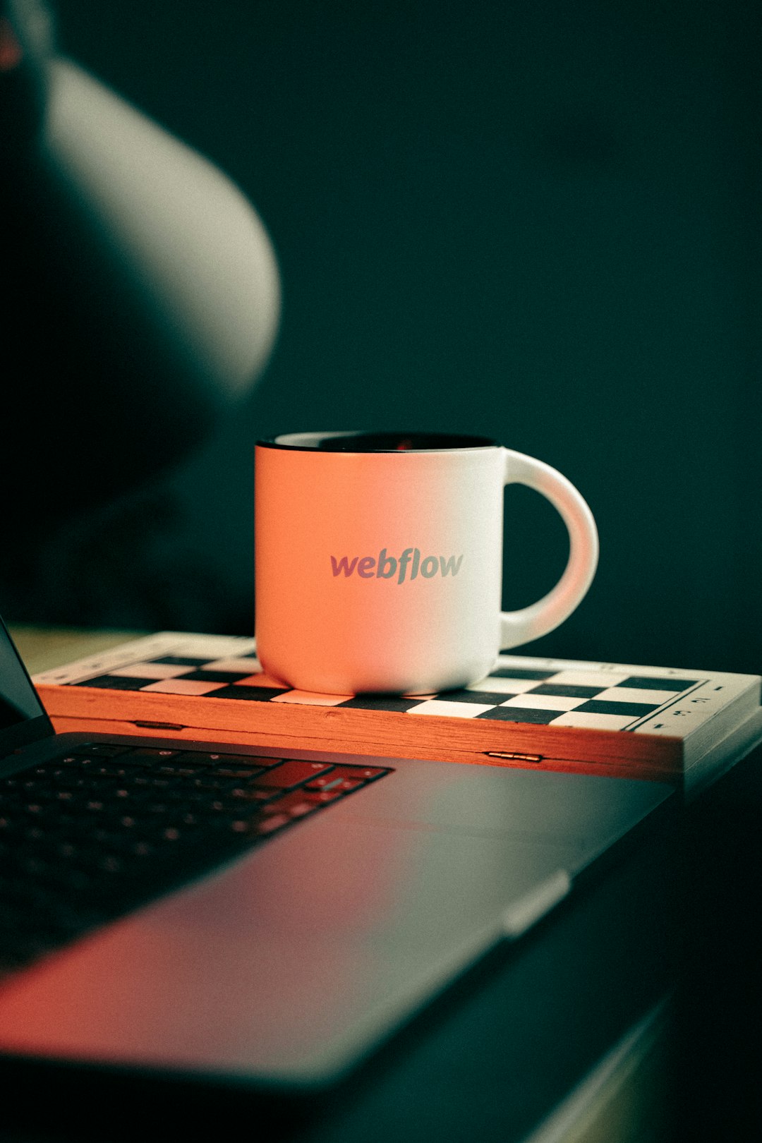 A coffee mug webflow logo on top of a chess and close to a MacBook Pro